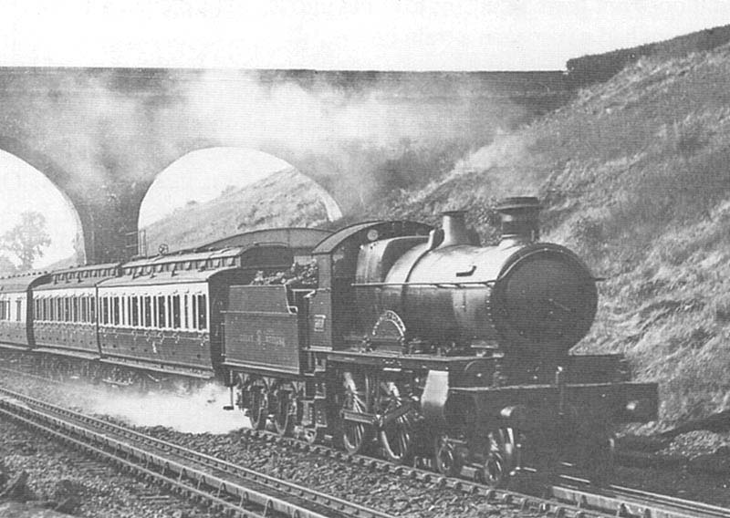 An unidentified GWR 4-4-0 County Class locomotive hauling a rake of mixed coaching stock passes over Rowington troughs on an up service
