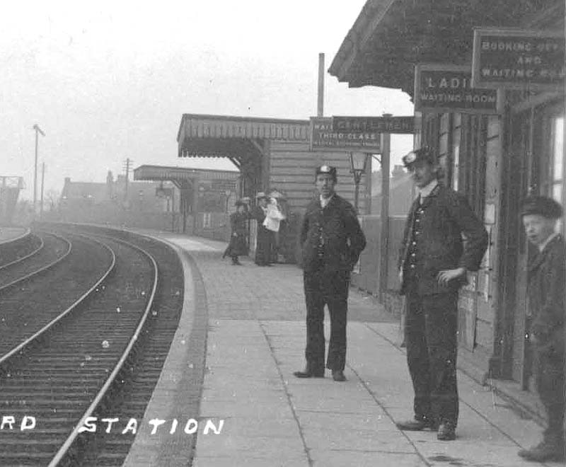 Close up showing three staff, the additional waiting rooms erected in the later 19th century and station signage