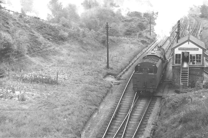 Ex-LMS 8F 2-8-0 No 48757 is running light engine and tender first as the signalman stretches out of the cabin to pass over the train staff circa 1955