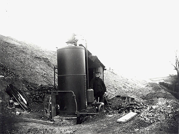 A navvy poses for the camera besides a pumping engine fitted with a vertical boiler at the foot of an embankment near Caves Inn, north of Newton