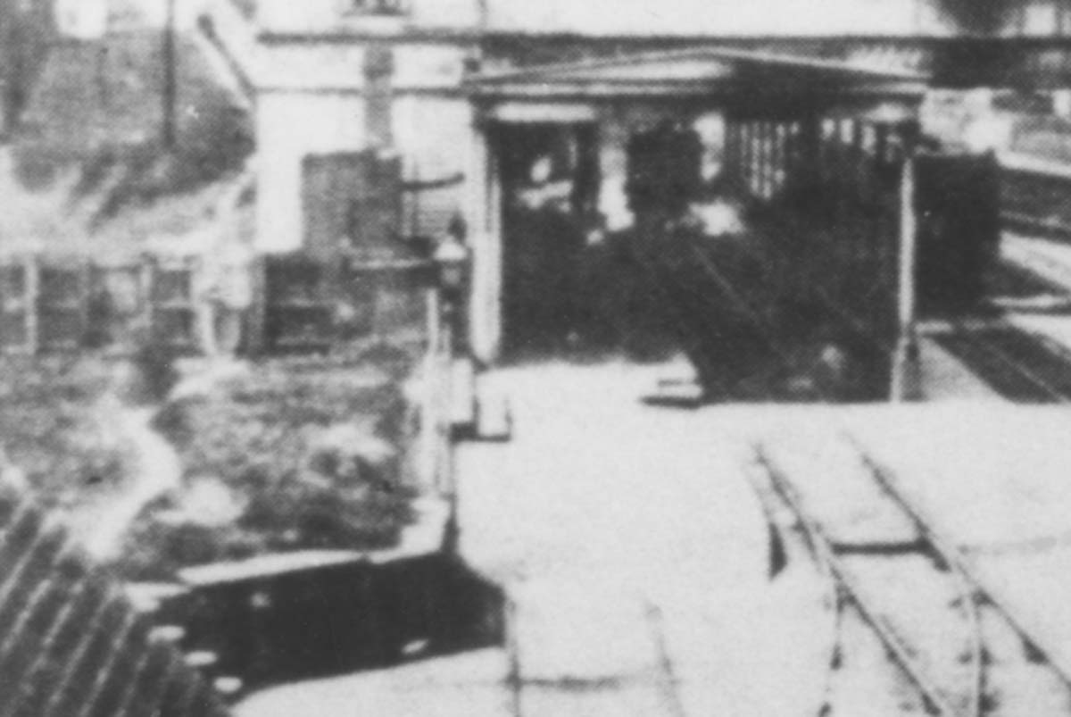 Close up of the down platform, train shed and passenger footbridge whilst a locomotive is seen underneath taking on water