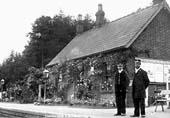 View of the simple station building bedecked with flowers with the Station Master and a porter posed on the platform